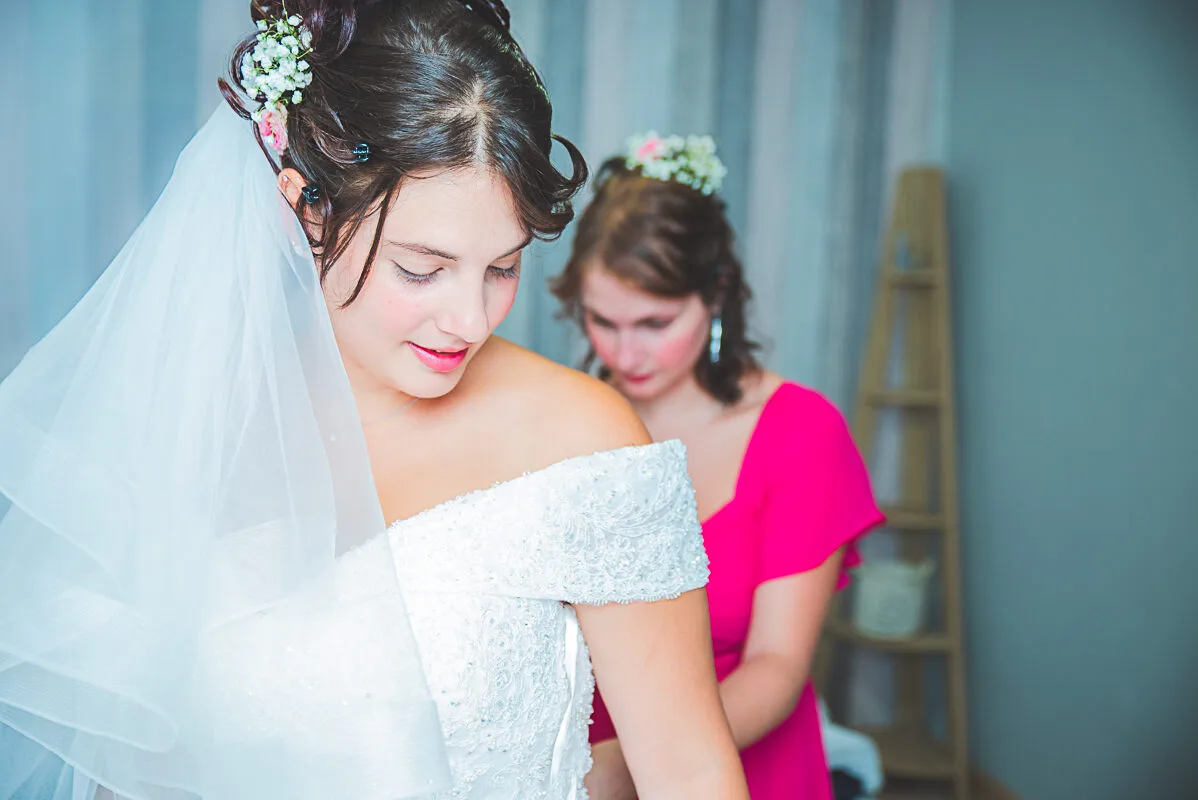 Préparatifs de la mariée entourée de sa soeur qui l'aide à fermer sa magnifique robe.